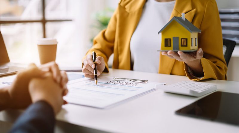 Image of someone sat across a desk from another person with a contract and holding a model home to support home building regulations article