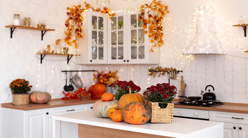 A bright, white kitchen decorated for fall with pumpkins, red mums, woven baskets, and warm string lights draped around cabinets and shelves.