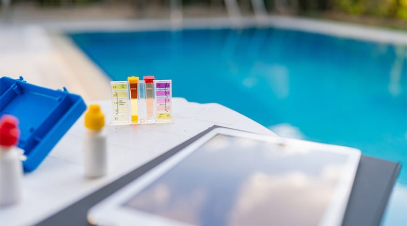 Pool water testing kit with color comparison vials and chemicals on a table beside a residential swimming pool.