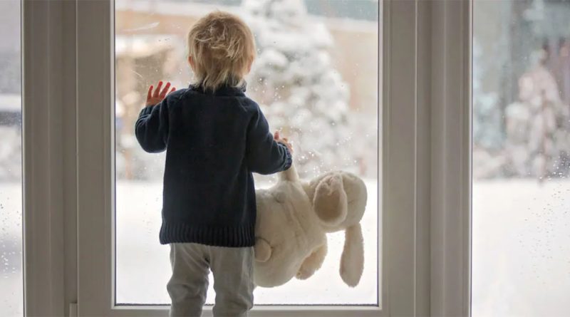 Young child standing indoors by a glass door, holding a stuffed animal and looking out at a snowy winter scene outside.