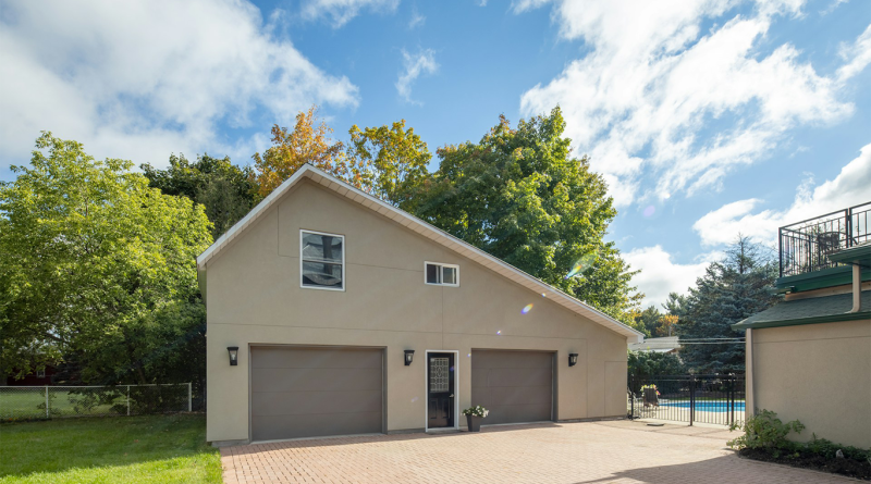 Beige detached two-car garage with pitched roof, double grey doors, and paved driveway under a blue sky, surrounded by trees and a fenced backyard with a pool.