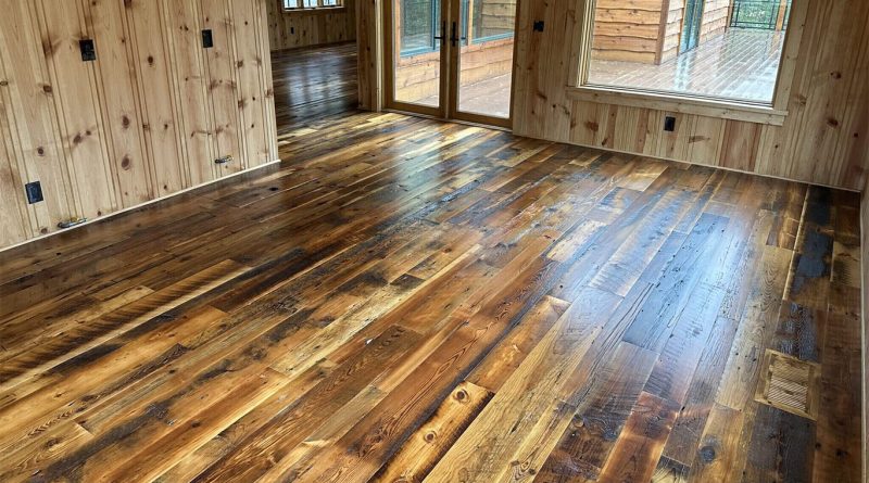 Interior room with wide-plank reclaimed wood flooring in varied natural tones, surrounded by wood-paneled walls and large windows overlooking a deck.